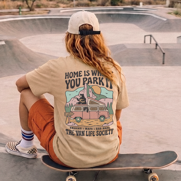 A person sitting on a skateboard at a concrete skatepark, viewed from behind, wearing a beige graphic t-shirt with a retro van illustration and the text 'HOME IS WHERE YOU PARK IT' and 'THE VAN LIFE SOCIETY'.