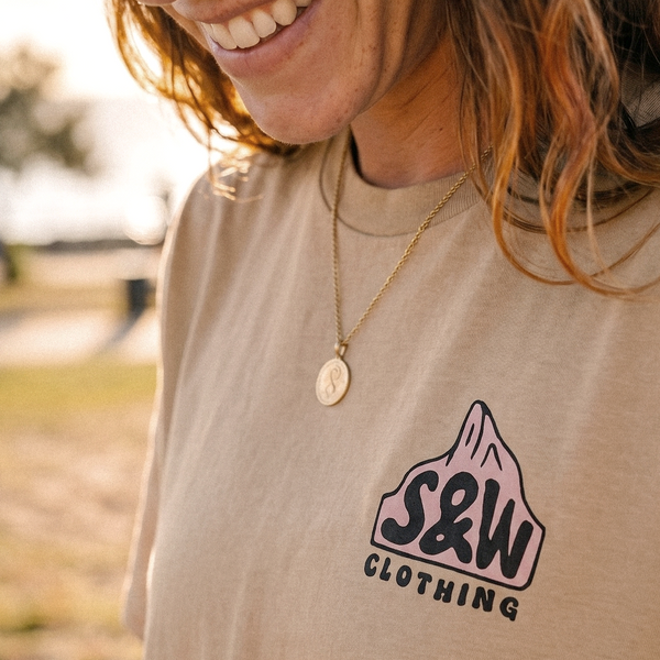 Close-up of a person wearing a beige graphic T-shirt with a pink mountain logo and the text 'S&W CLOTHING' on the chest, photographed outdoors with warm natural light.