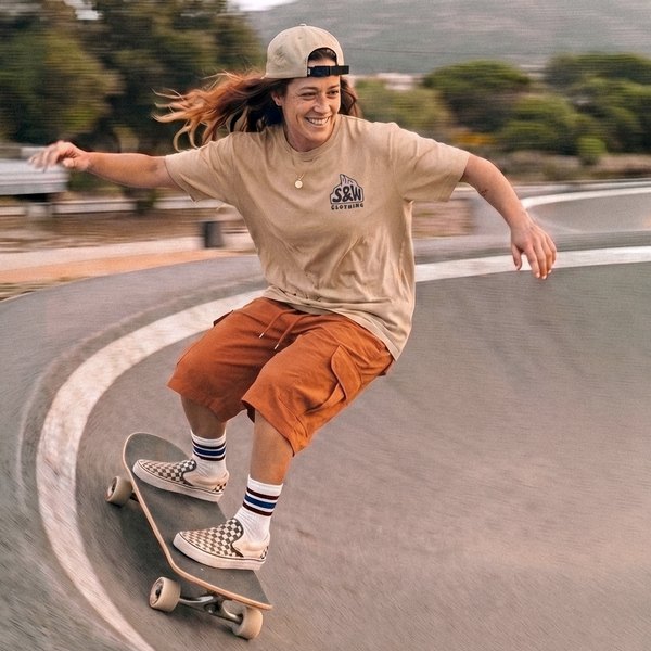 A person skateboarding in a curved outdoor ramp, wearing a beige t-shirt with a visible 'The Society' logo, rust-orange cargo shorts, striped socks, and checkered Vans slip-on shoes, with a beige cap backwards.