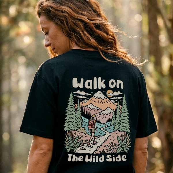A woman shown from behind wearing a black graphic t-shirt in an outdoor forest setting. The back of the shirt features a colorful illustration of a mountain, pine trees, and a hiking trail, with the text 'Walk on The Wild Side' printed in white.