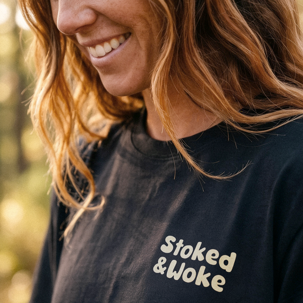 A close-up lifestyle shot of a woman outdoors wearing a black crewneck t-shirt with the words 'Stoked & Woke' printed in a light beige retro font on the chest.