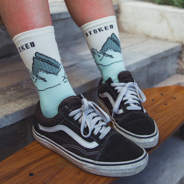 Person wearing 'Stoked' socks with mountain design and black Vans shoes on a wooden surface.