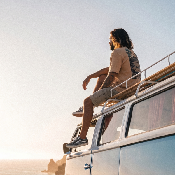 A man with long dark hair sits on the roof of a light blue and white vintage camper van, looking out toward the ocean under a clear sky. He is wearing a beige t-shirt, beige shorts, and black sneakers.