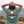 A person viewed from behind wearing an olive green t-shirt with a large graphic on the back showing mountains, a sun, and the text 'NATURES BALANCE'. The background is a blurred outdoor landscape with mountains and trees.