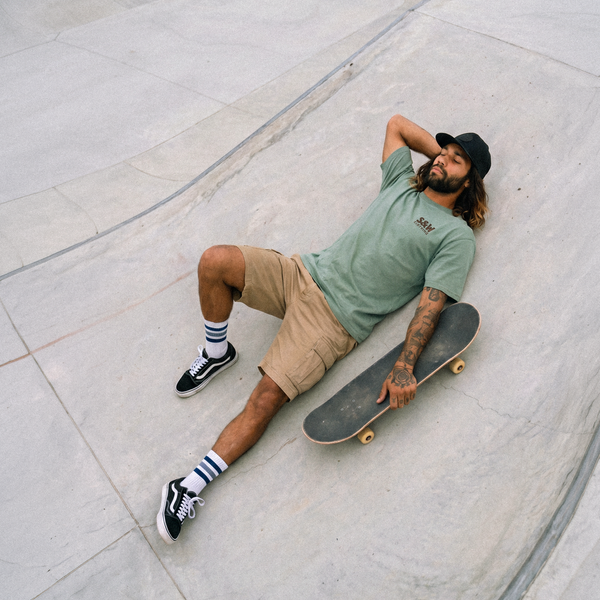 A man with a beard lies on a concrete skatepark ramp holding a skateboard, wearing a sage green t-shirt, khaki shorts, black and white socks, and black Vans sneakers.